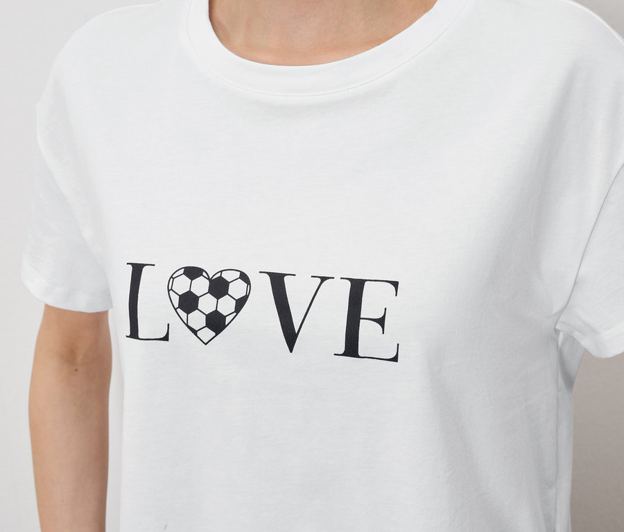 Közeli kép egy személyről, aki egy fehér pólót visel, amin a "LOVE" felirat látható, ahol az "O" betűt egy futball-labda alakú szív helyettesíti.