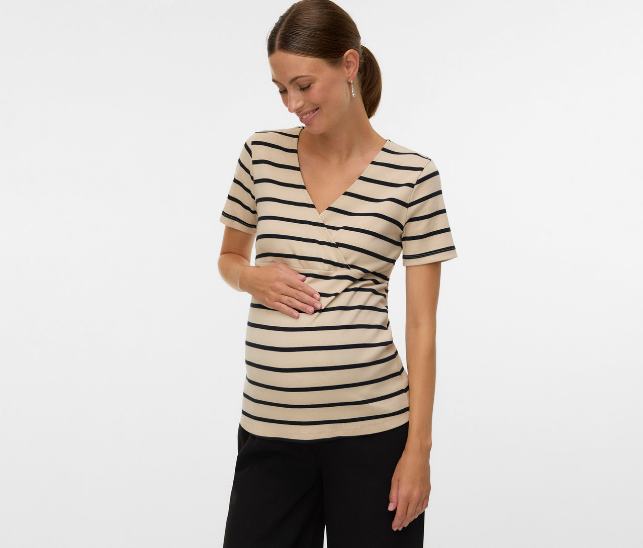 A lófarokba kötött hajú nő egy csíkos MAMALICIOUS kismamapólót »MLLANE TESS« és fekete nadrágot visel, miközben a hasát érinti.