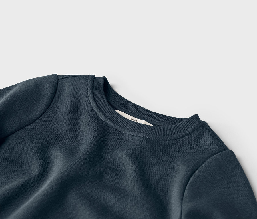 Egy sötétkék, kerek nyakú pulóver részletgazdag nézete, a gallérján a Tchibo márka címkéjével.