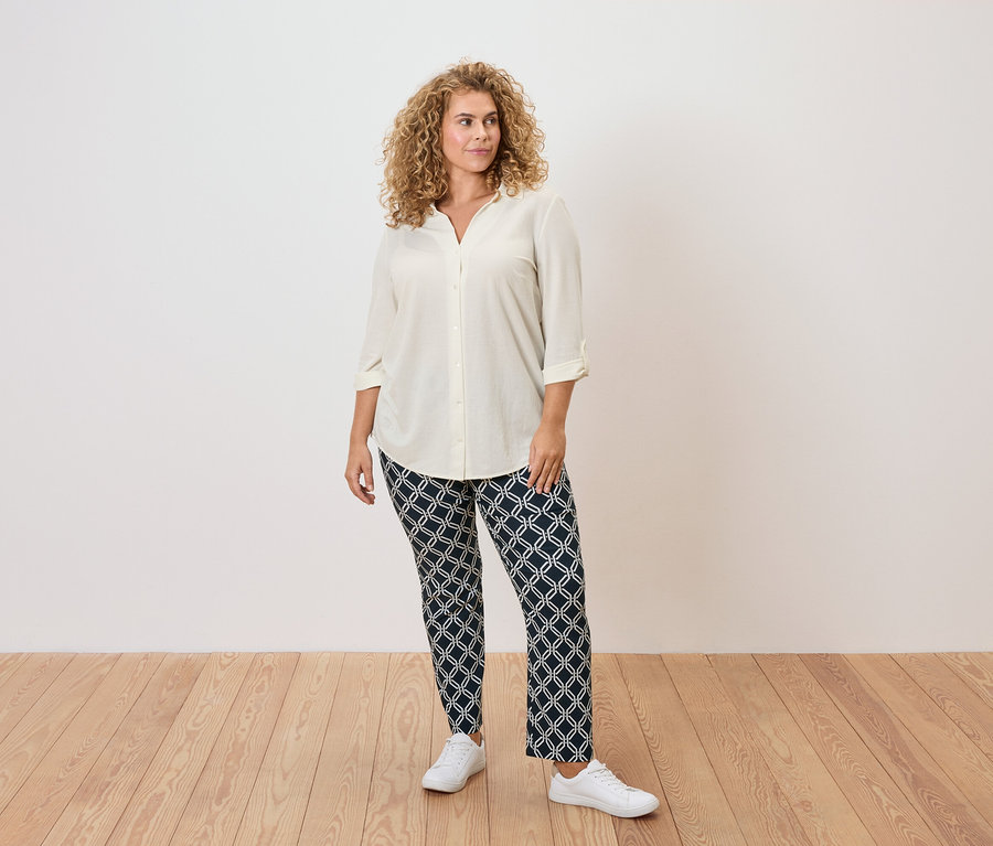 A göndör hajú nő krémszínű blúzt, mintás nadrágot és fehér tornacipőt visel.