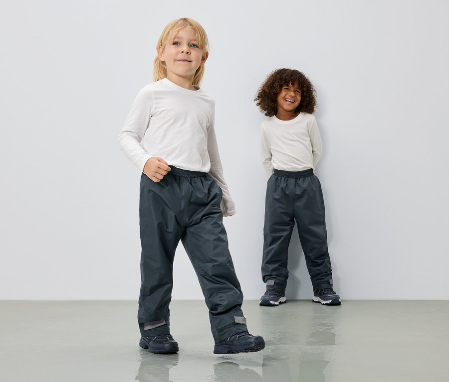 Két gyerek hosszú, szürke esőnadrágot és hosszú ujjú, fehér felsőt visel egy fehér fal előtt.