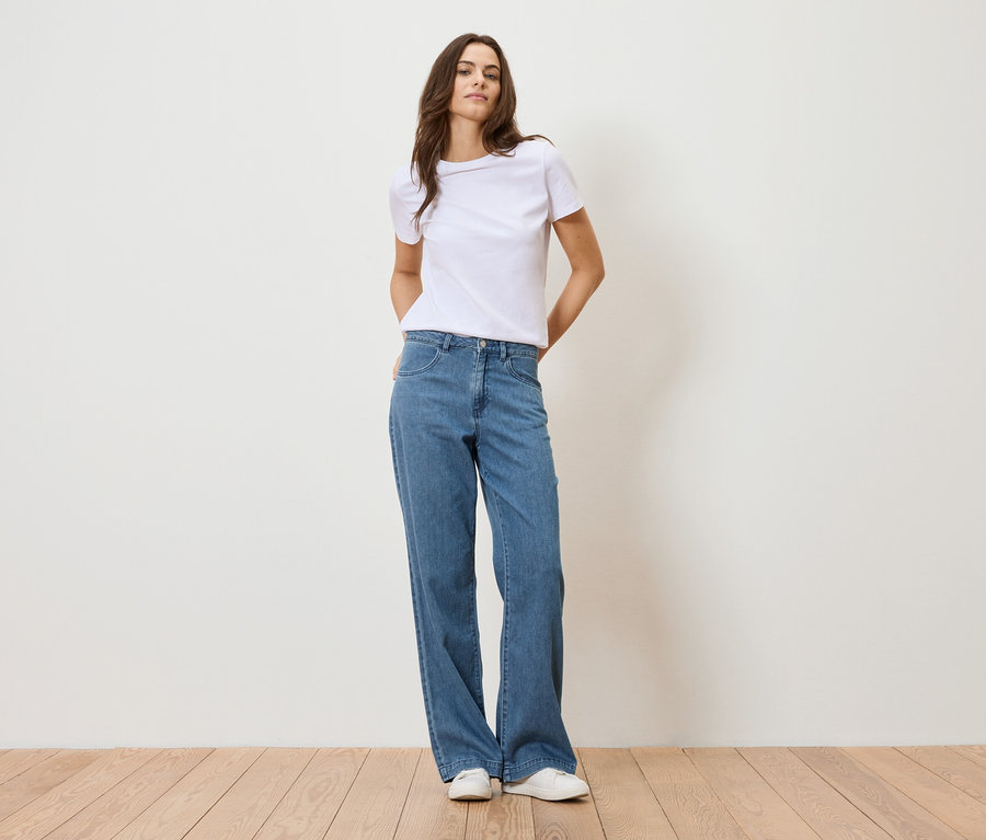 Nő áll egy fehér fal előtt, fehér pólót és kék farmert visel.