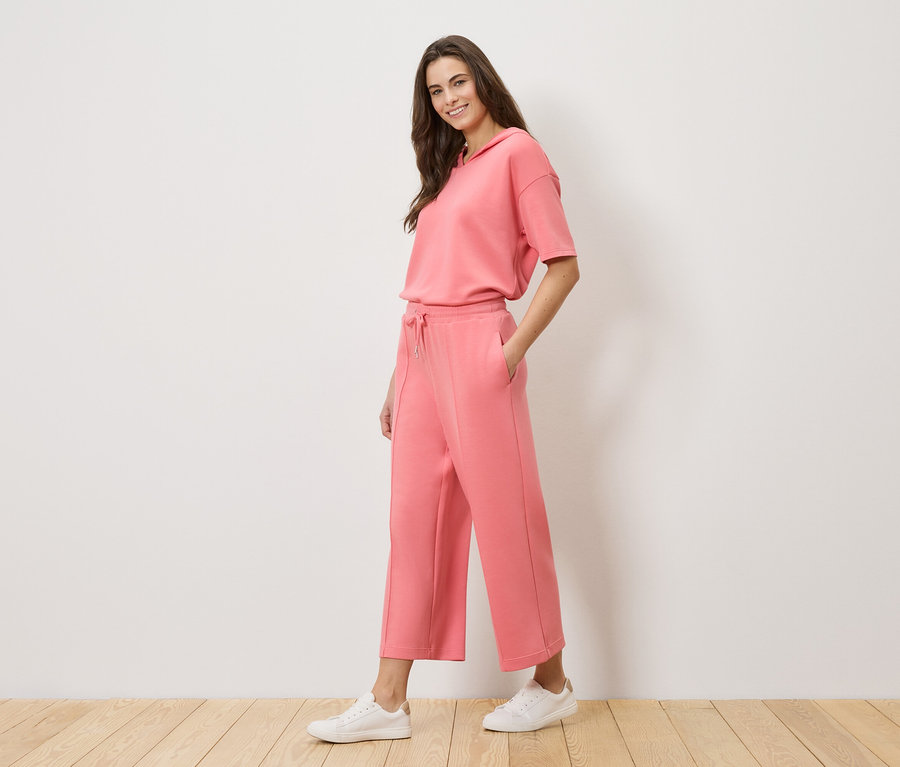 Nő korall színű tréningruhát visel kapucnis felsővel és culotte nadrággal, valamint fehér tornacipőt.