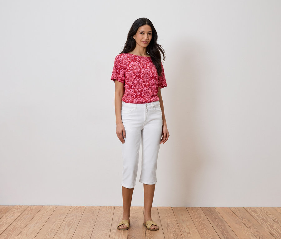 Nő áll a fal előtt, piros felsőt és fehér nadrágot visel.