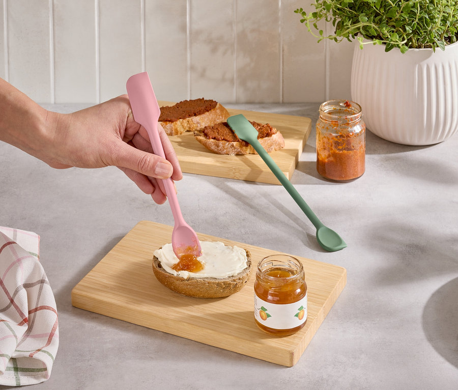 Egy kéz rózsaszín spatulával ken lekvárt egy zsemlére. Mellette egy zöld spatula és két üveg lekvár áll.