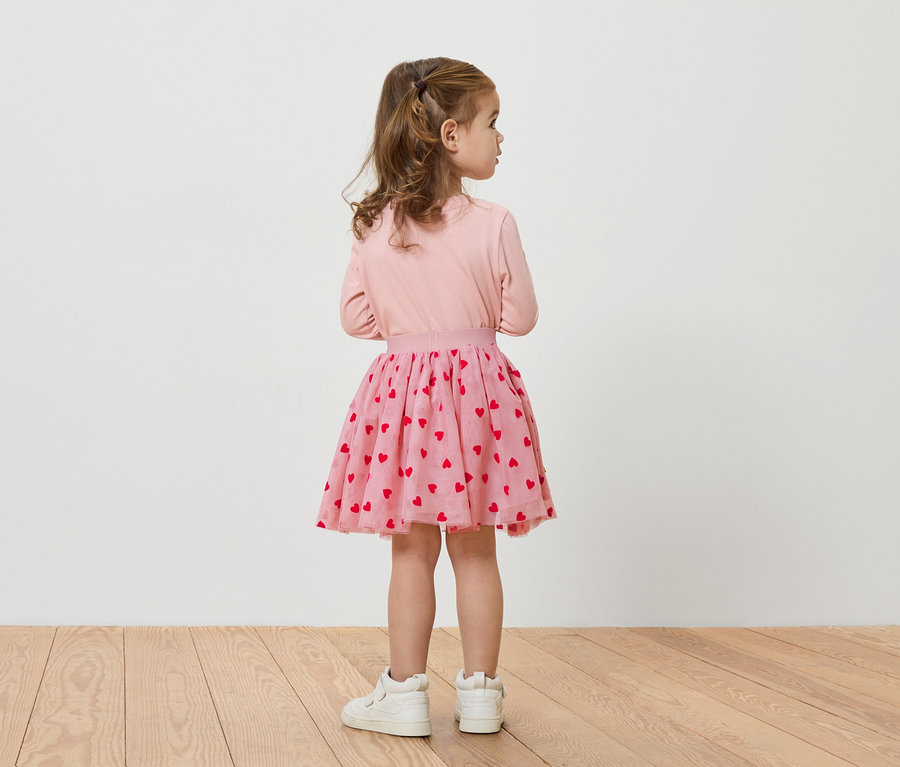 Hátulról látható kislány rózsaszín felsőben, piros szíves szoknyában és fehér cipőben.