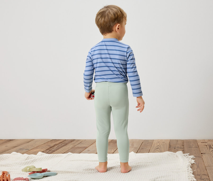 Egy gyermek áll háttal a kamerának, kék csíkos inget és világoszöld leggingset visel.