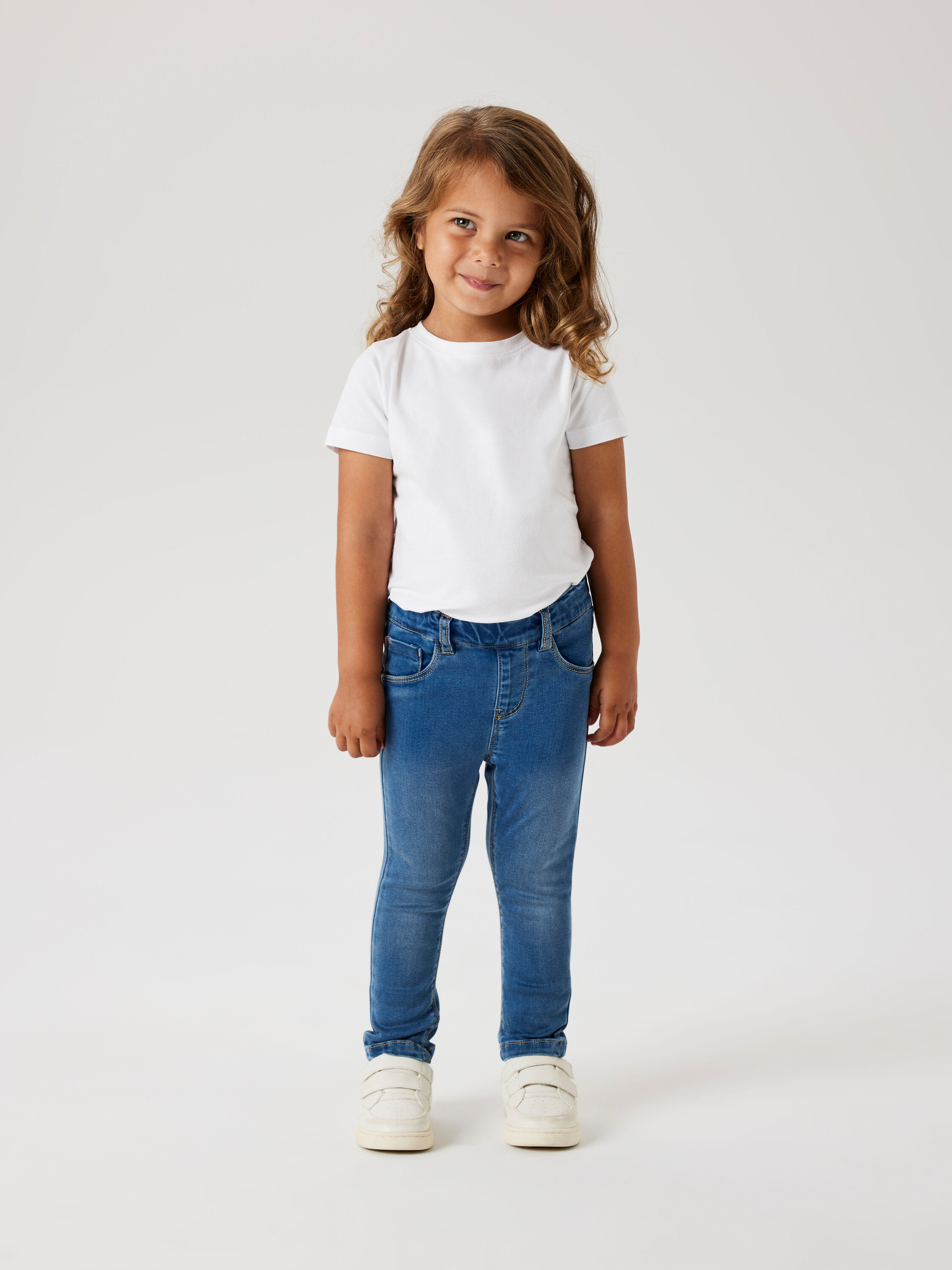 Göndör hajú lány fehér pólót, kék farmert és fehér cipőt visel.