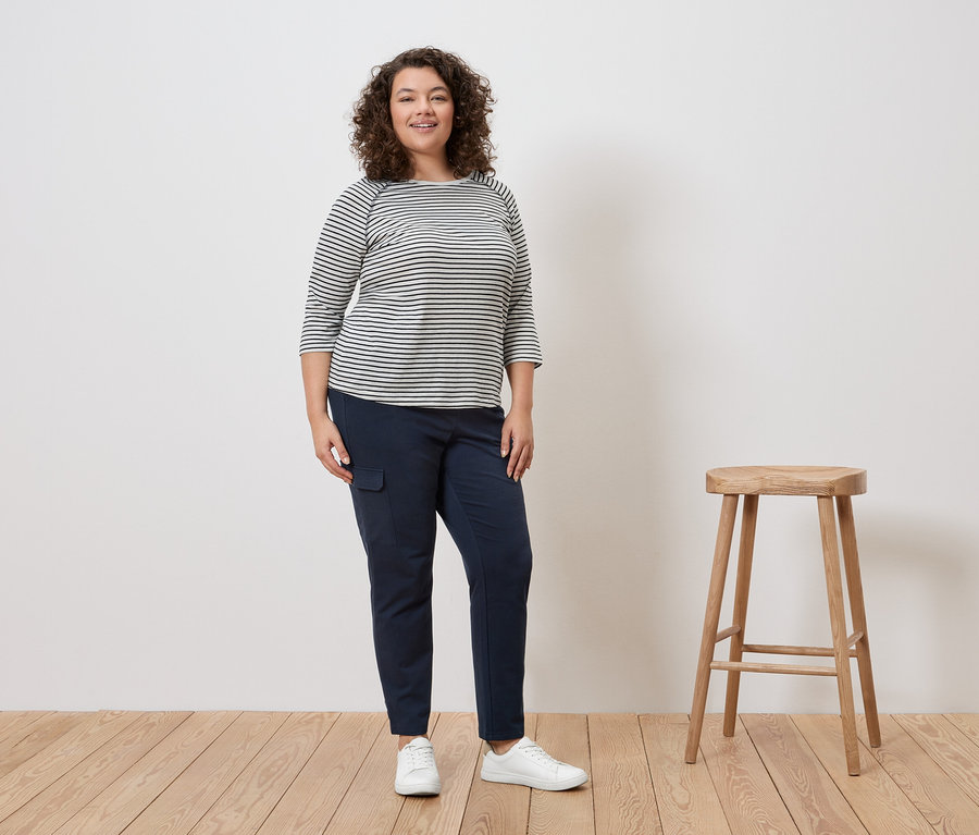 Göndör hajú nő áll csíkos felsőben, kék nadrágban és fehér tornacipőben egy zsámoly mellett.