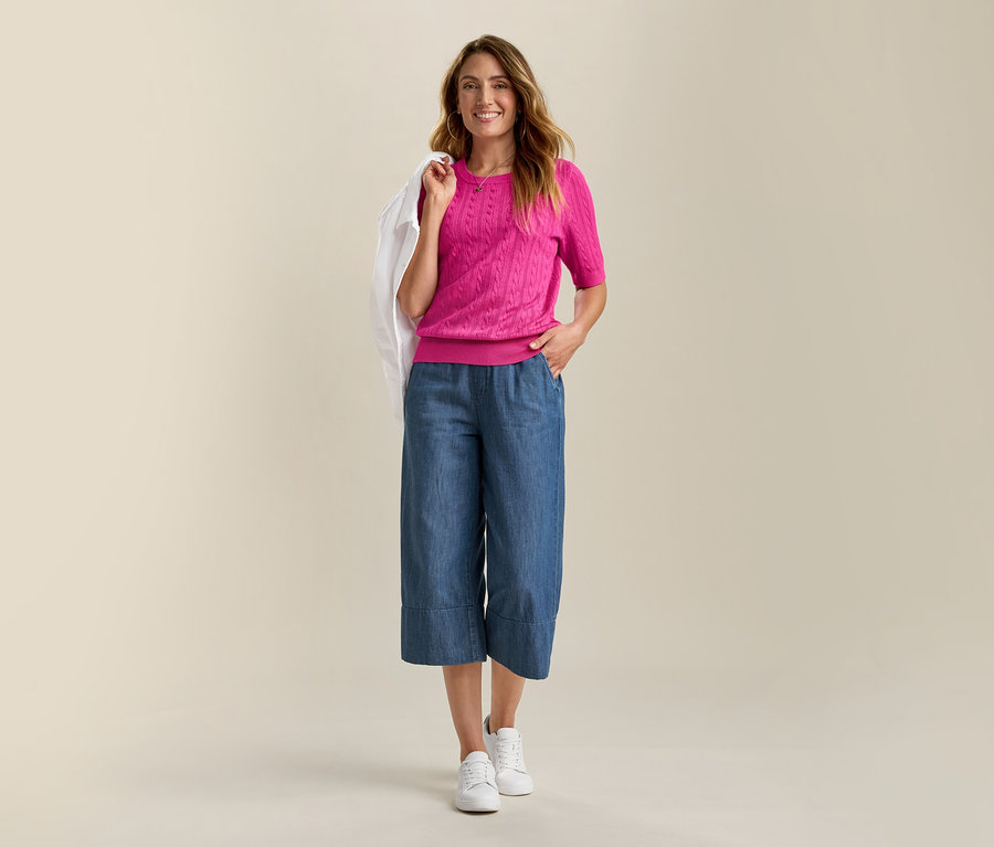 Mosolygó nő farmerdzsekiben és rózsaszín, női finomkötésű, fonott mintás pulóverben, kék culotte nadrágot és fehér tornacipőt visel.