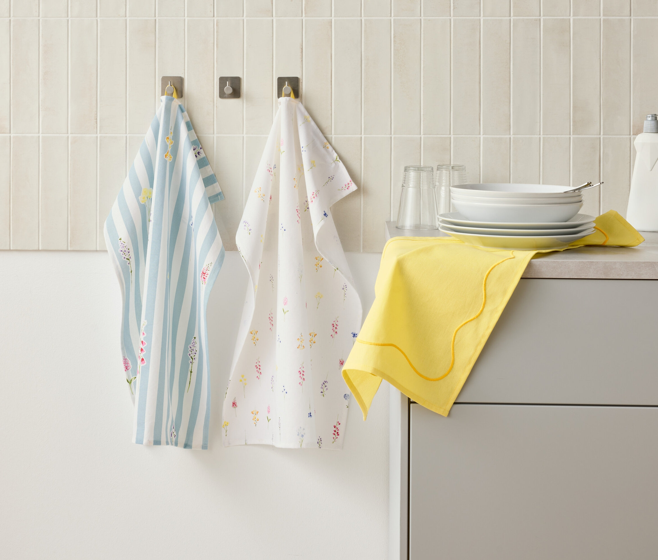 Két konyharuha lóg kampókon a falon egy konyhapult fölött, amelyen edények, poharak, mosogatószeres flakon és egy sárga kendő található.