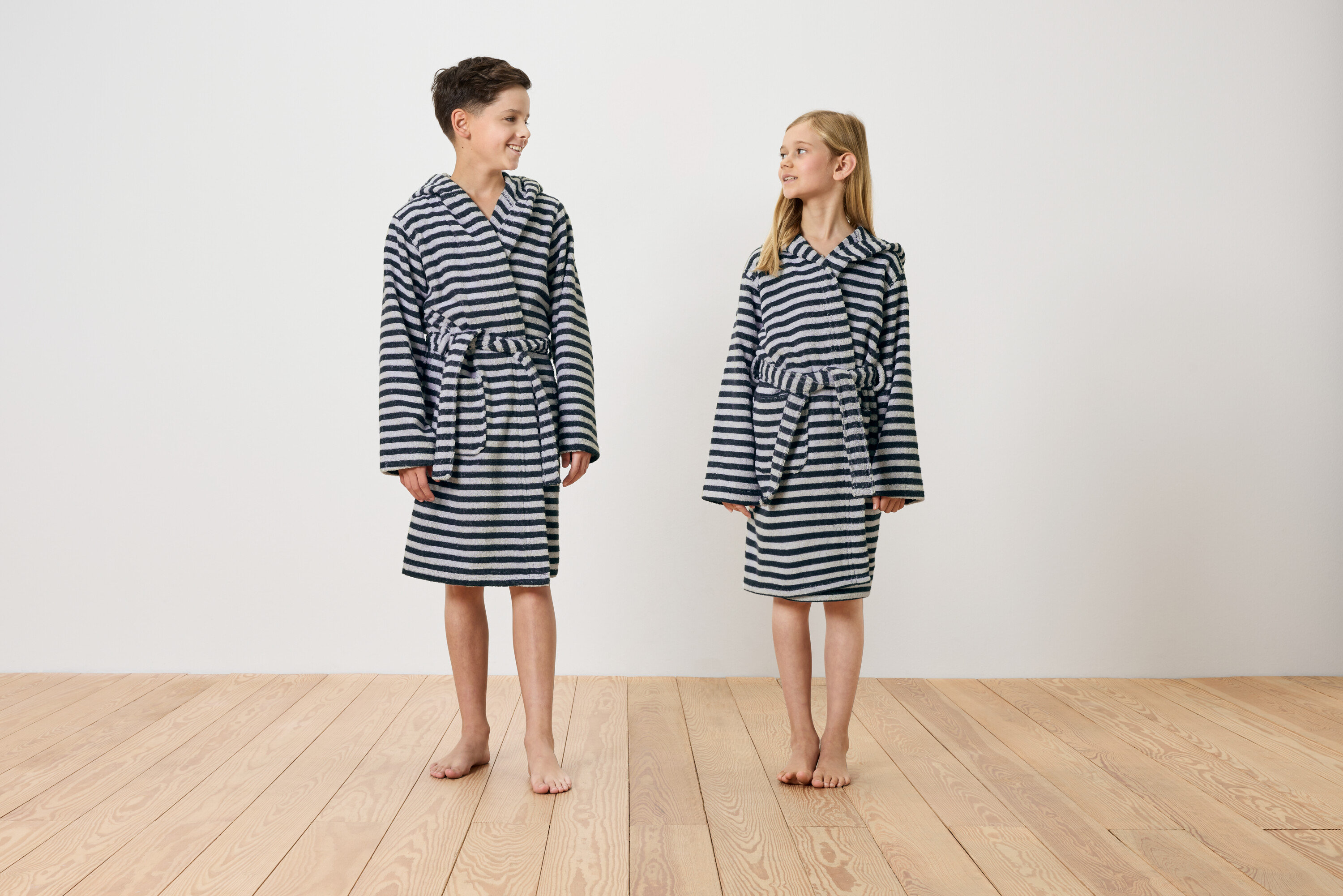 Egy fiú és egy lány mezítláb állnak egy fapadlón, csíkos, kapucnis köntösben.