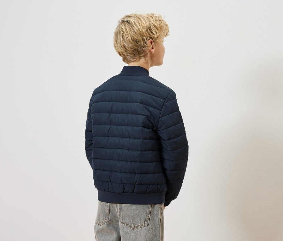 Hátulról látható egy szőke gyermek tengerészkék steppelt kabátban és szürke farmerben, fehér háttér előtt.