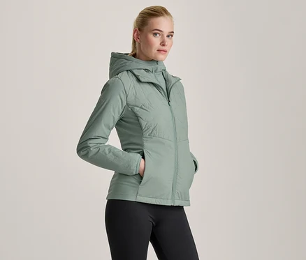 Világoszöld kapucnis kabátot és fekete leggingset viselő nő, keze a kabát zsebében.