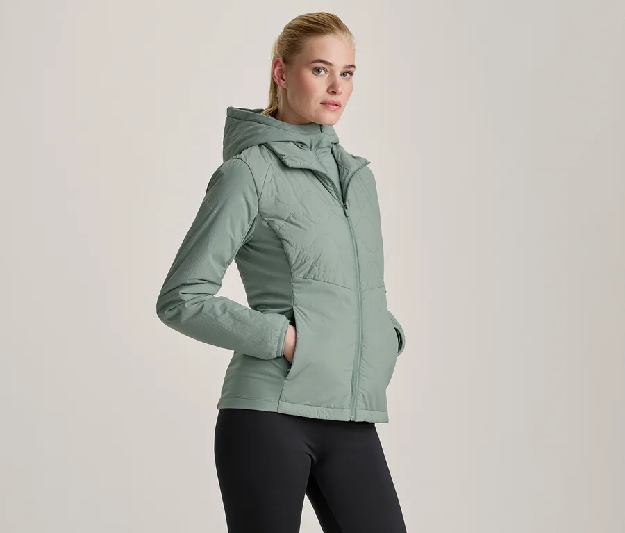 Világoszöld kapucnis kabátot és fekete leggingset viselő nő, keze a kabát zsebében.