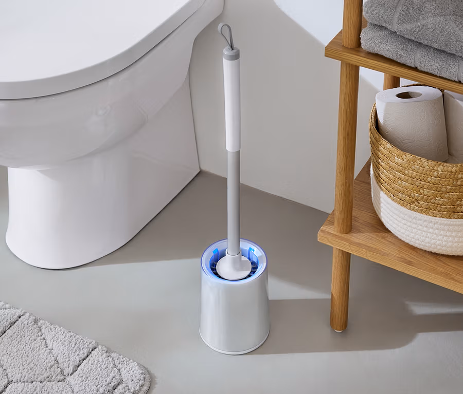 A fürdőszobában egy fehér WC áll egy kék fényű WC-kefe és egy törölközőkkel és WC-papírral ellátott fa polc mellett.