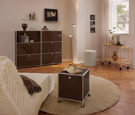 Modern nappali bézs kanapéval, nagy barna fém »CN3« komóddal 3 lehajtható rekesszel, kis barna fém »CN3« gurulós komóddal 2 lehajtható rekesszel kerek szőnyegen, buklé ülőkével és fa tálalókocsival.