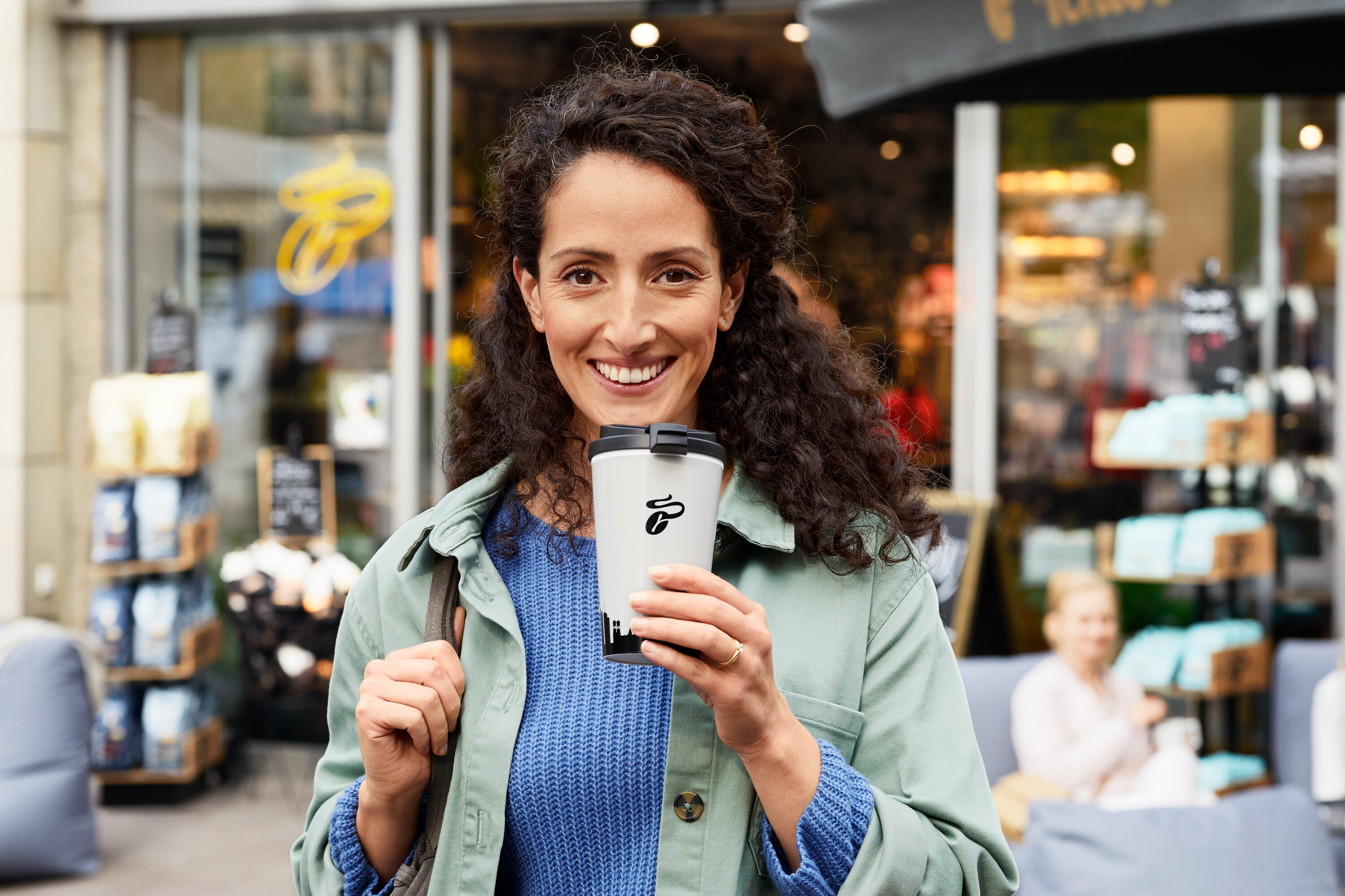 Mosolygó nő tart egy fehér Tchibo termobögrét egy Tchibo üzlet előtt.