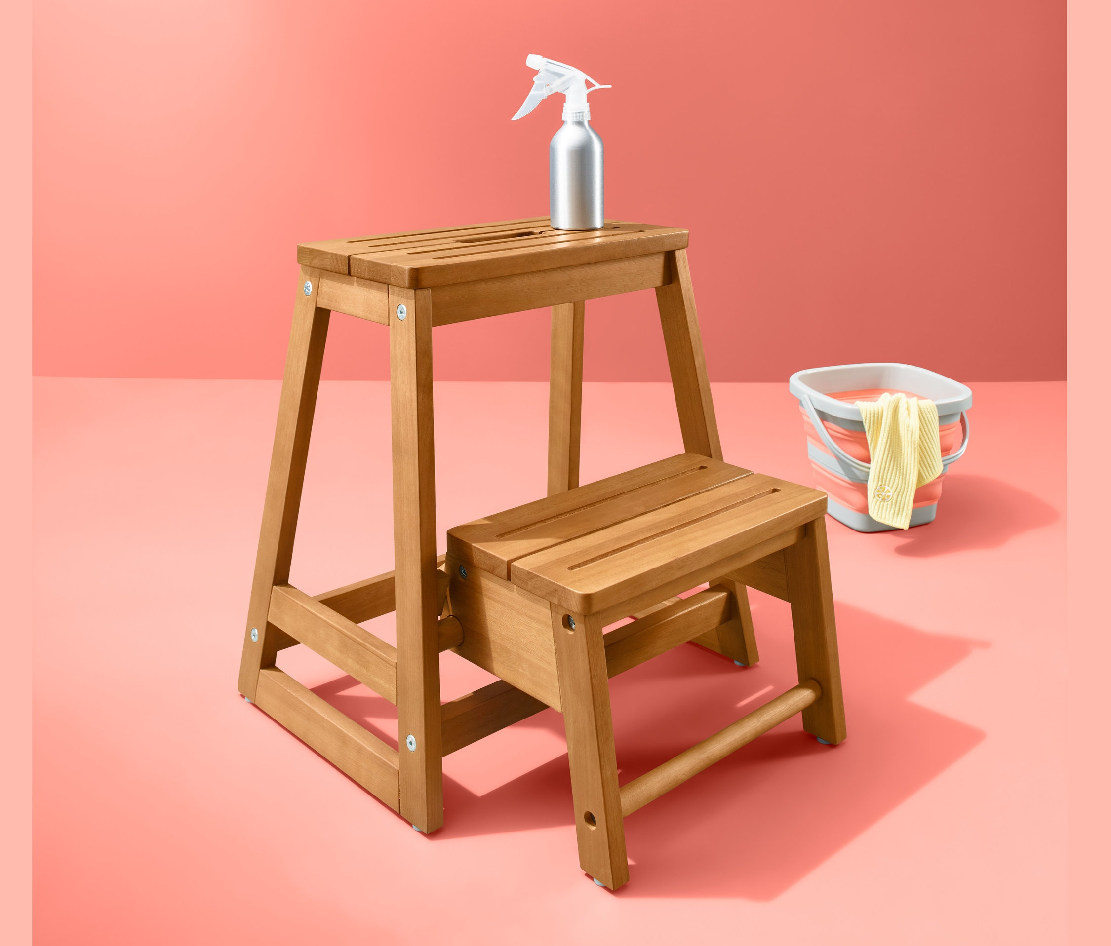Egy fa összecsukható fellépő, egy szórófejes flakon és egy vödör korall színű háttér előtt.