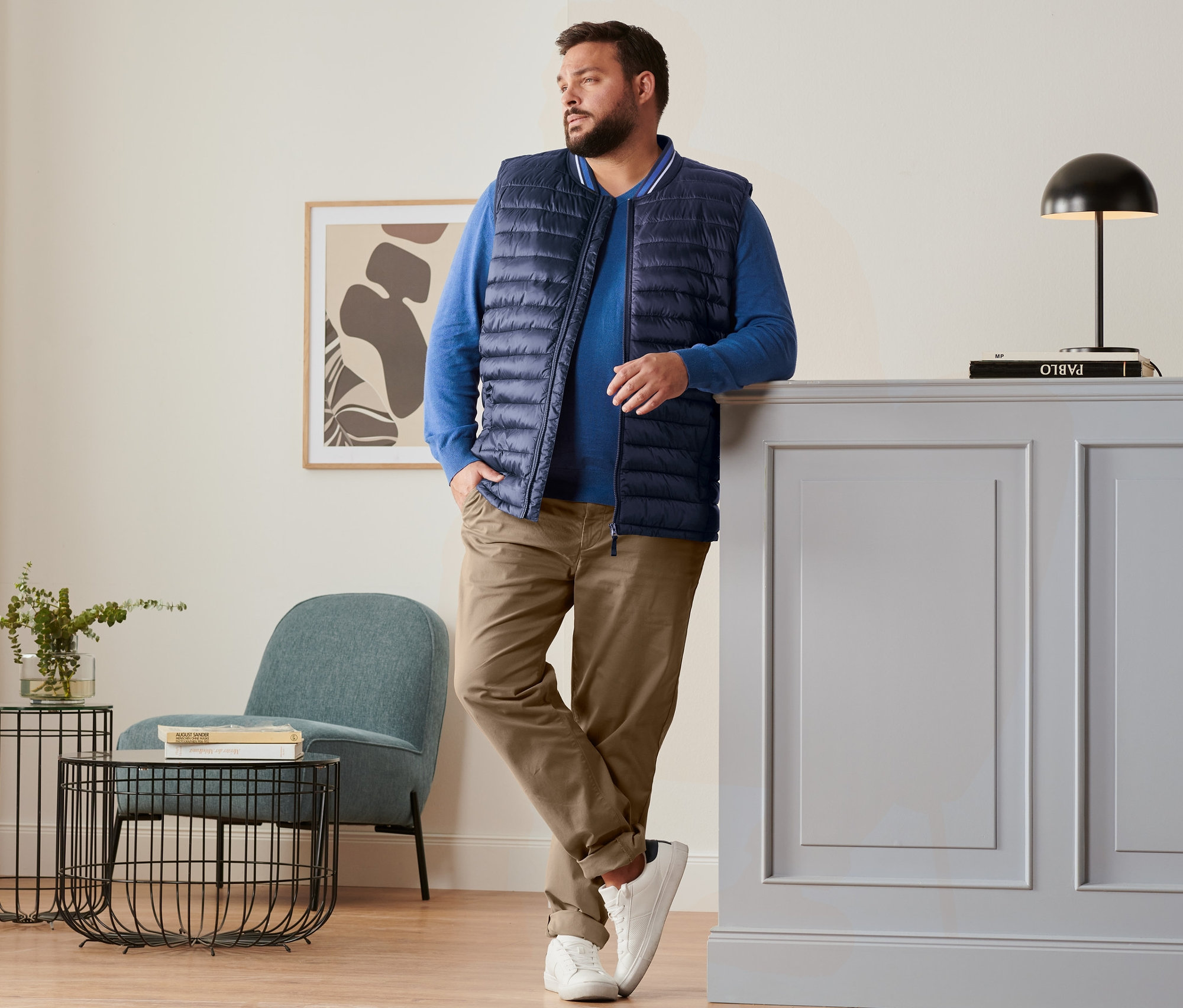 Férfi támaszkodik a szekrénynek, sötétkék steppelt mellényt, kék lenvászon pulóvert, barna chino nadrágot és fehér sneaker cipőt visel.