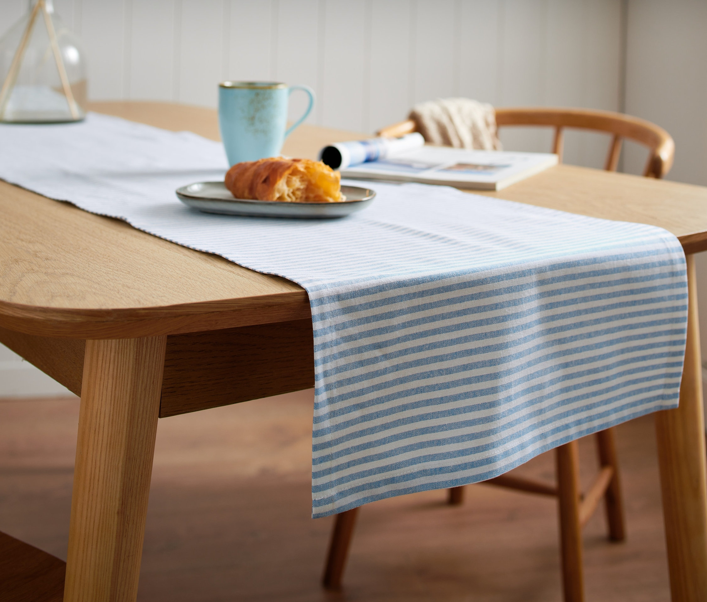 A fa asztalon egy kék-fehér csíkos asztali futó található. Rajta egy tányér péksütemény és egy kék bögre.