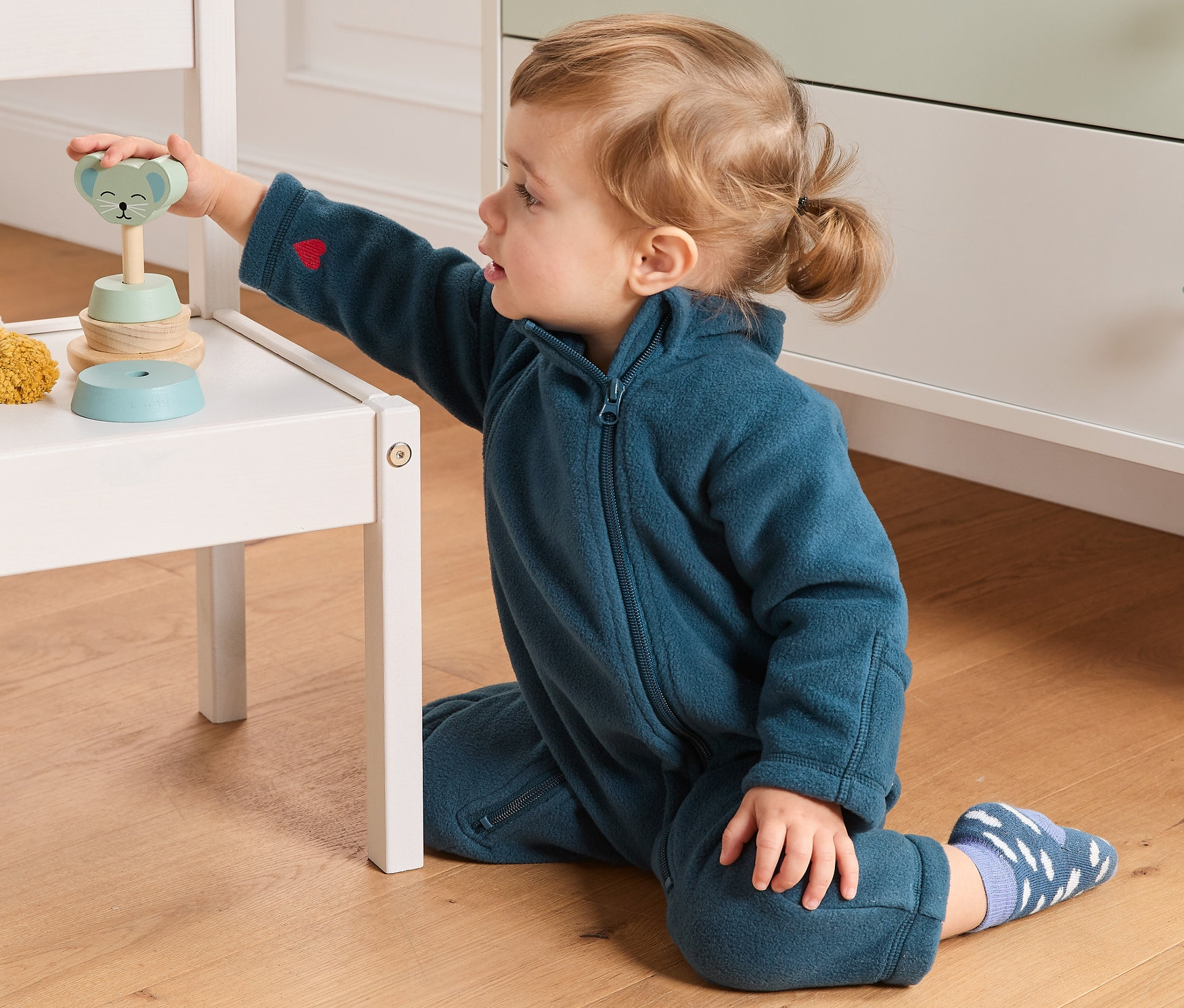 Kisgyermek játszik egy fa játékkal egy fehér széken, sötétkék színű polár kezeslábast visel.