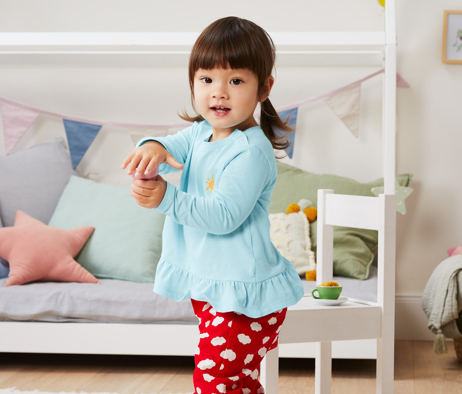 Egy kislány áll a gyerekszobában, világoskék felsőt és piros, fehér felhős nadrágot visel. A szettben lévő 3 kislány felső szettben, piros/sárga/világoskék színben van.