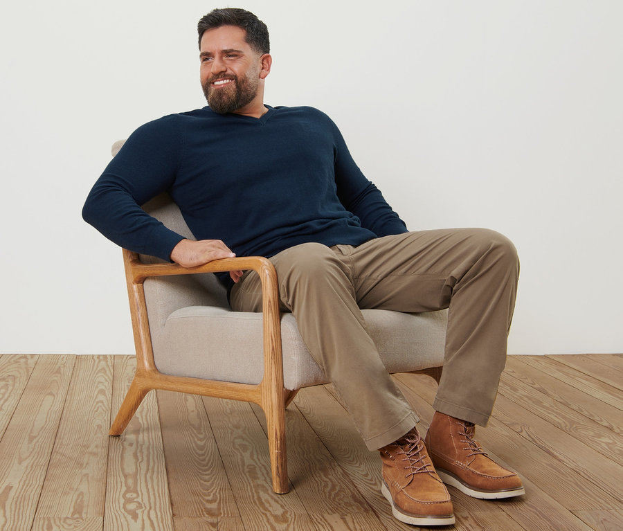 Egy férfi ül egy fotelben, homokszínű chino nadrágot és tengerészkék V-nyakú pulóvert visel.