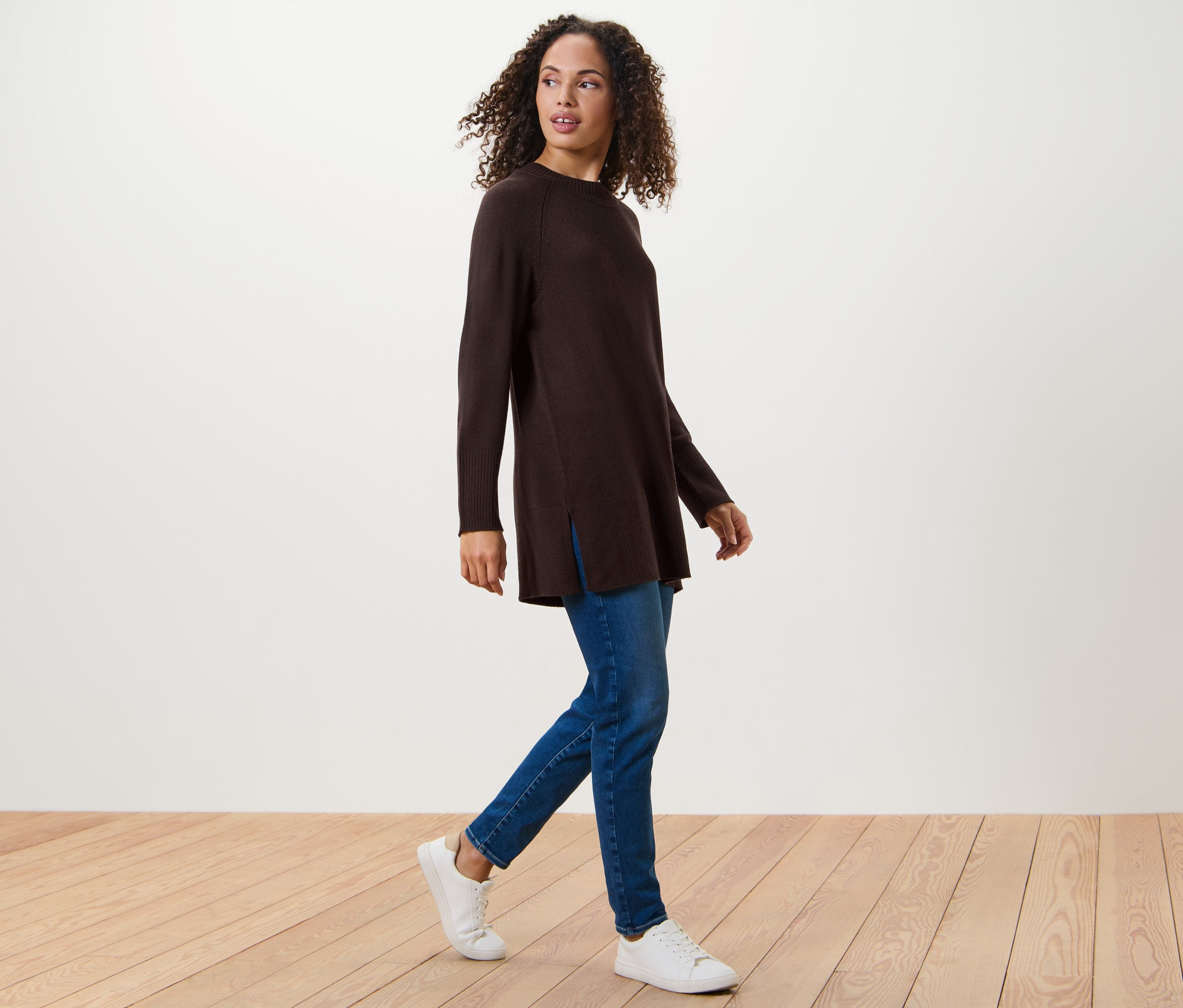 Nő sétál, sötétbarna hosszú kötött pulóvert, farmert és tornacipőt visel.