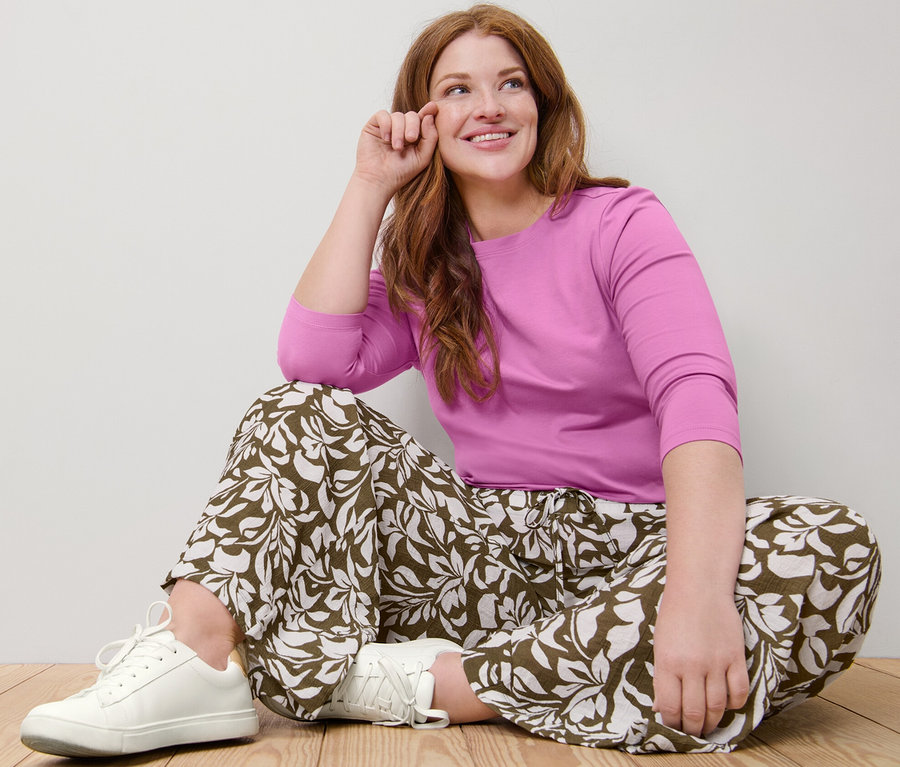 Nő ül a padlón rózsaszín, 3/4-es ujjú felsőben és nyomott mintás nadrágban.