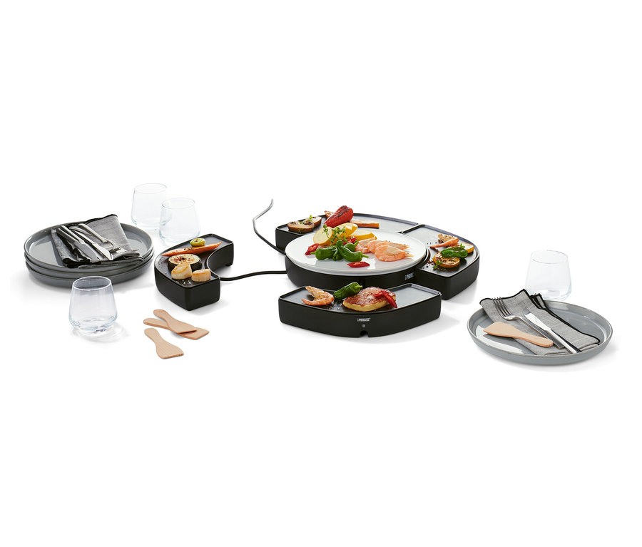 Fekete »Dinner4All« asztali grill ételekkel a grillfelületen és egy tányéron. Körülötte tányérok evőeszközökkel, poharak és fa spatulák.