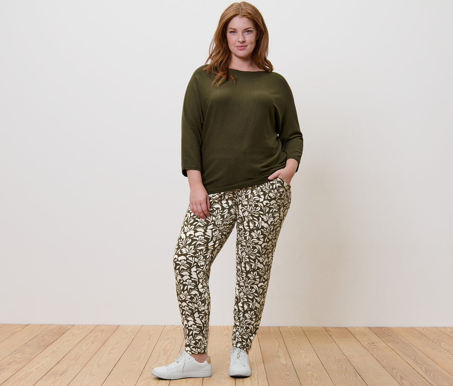 Egy nő áll zöld finomkötésű pulóvert és mintás jerseynadrágot viselve.