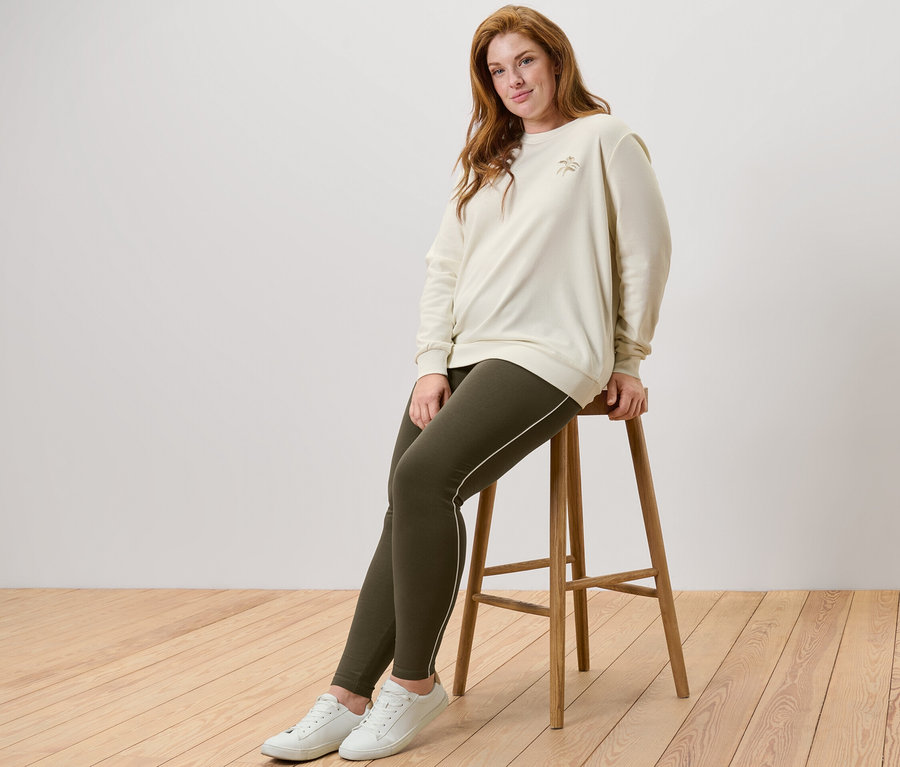 Nő ül egy széken, krémszínű, hímzett szabadidőfelsőt és leggings paszpólt visel.