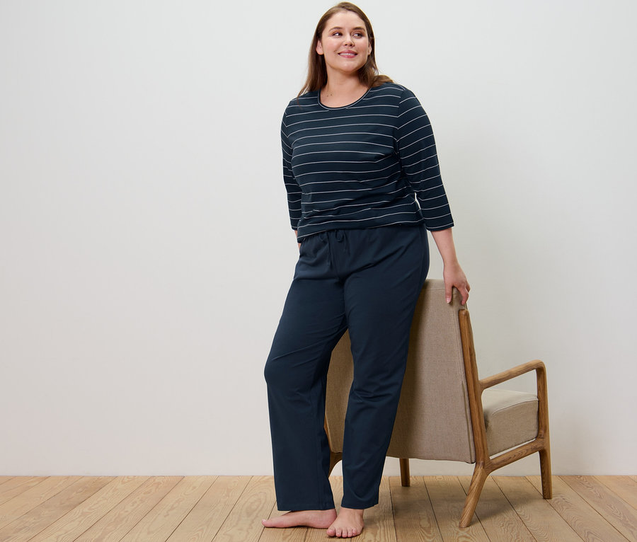 Nő sötétkék csíkos pizsamában áll egy szék mellett.