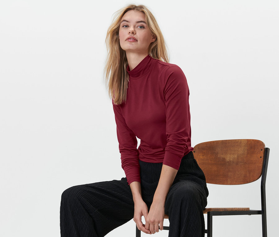 Egy szőke nő ül egy széken, piros állógalléros felsőt és fekete nadrágot visel.