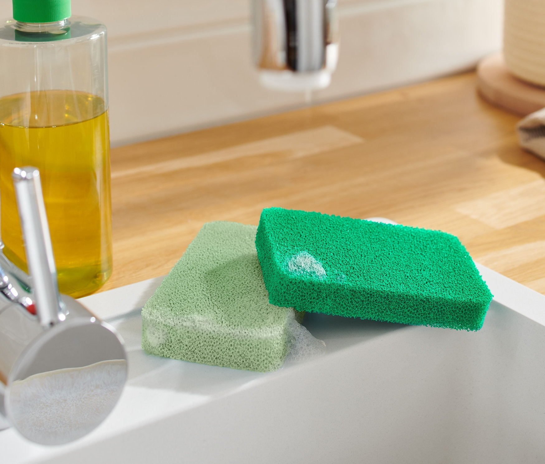 Két szilikon szivacs egy fehér mosogatótálcában, egy csaptelep és egy tisztítószeres flakon mellett.