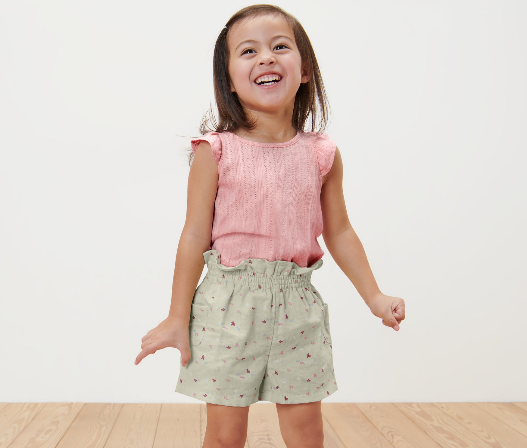 Egy mosolygós kislány áll, és bő gyerek rövidnadrágot és egy rózsaszín felsőt visel.