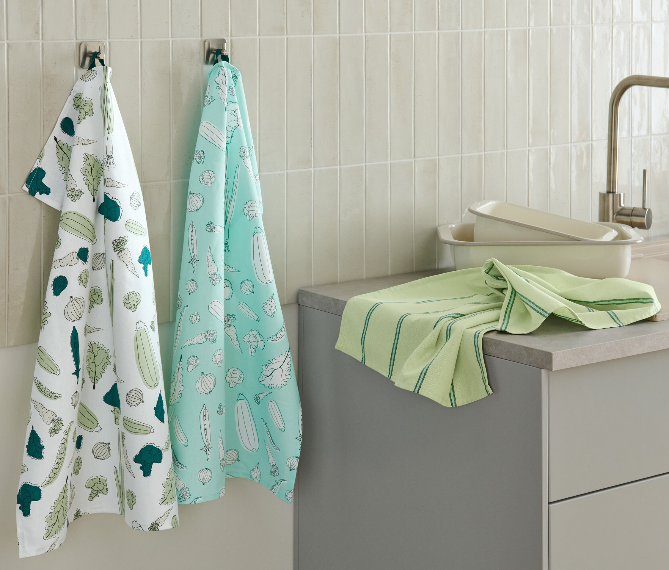 Két zöldségmintás konyharuha lóg a falon, egy másik zöld konyharuha a konyhapulton.