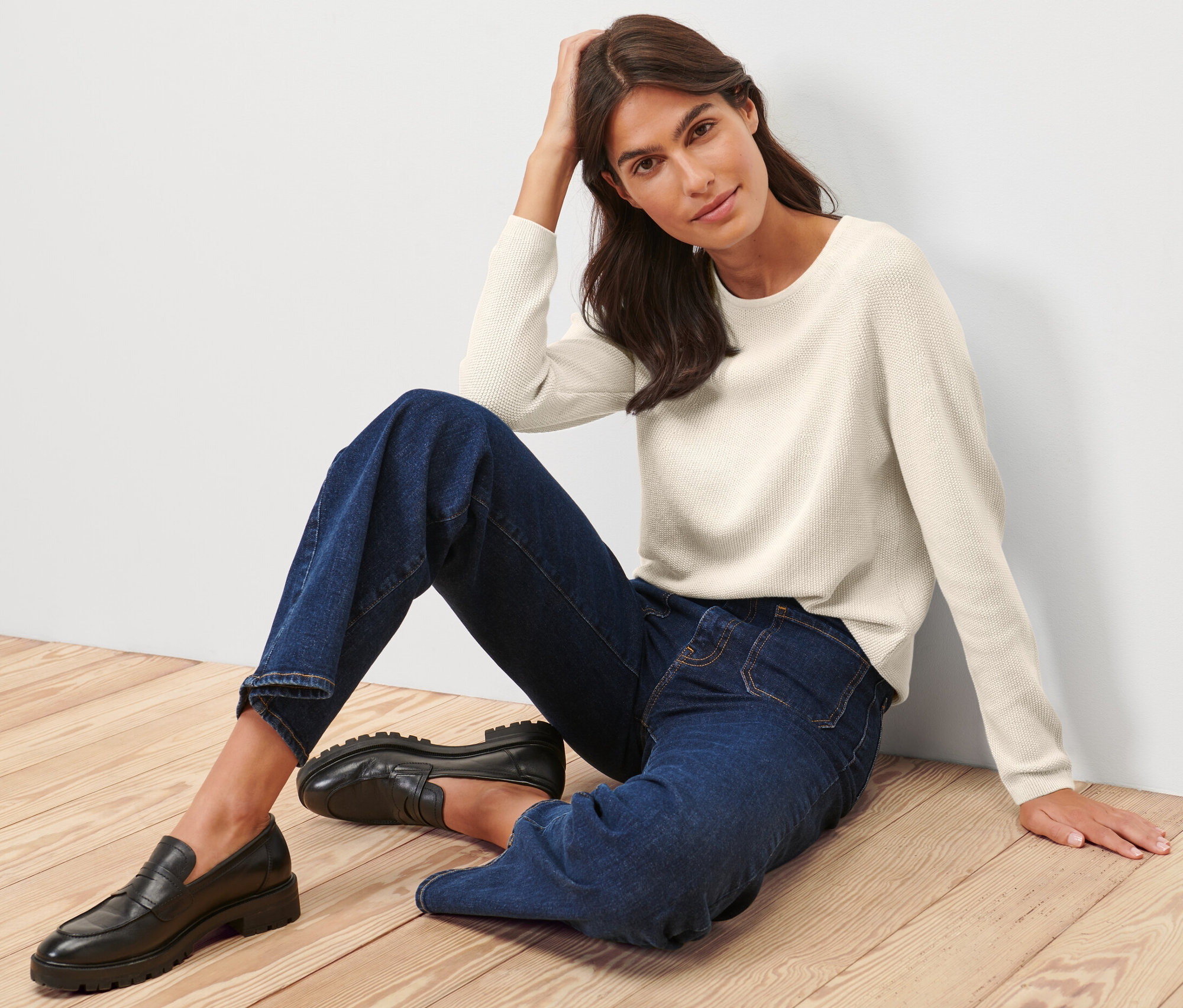 Nő ül a fapadlón, krémfehér kötött pulóvert visel.