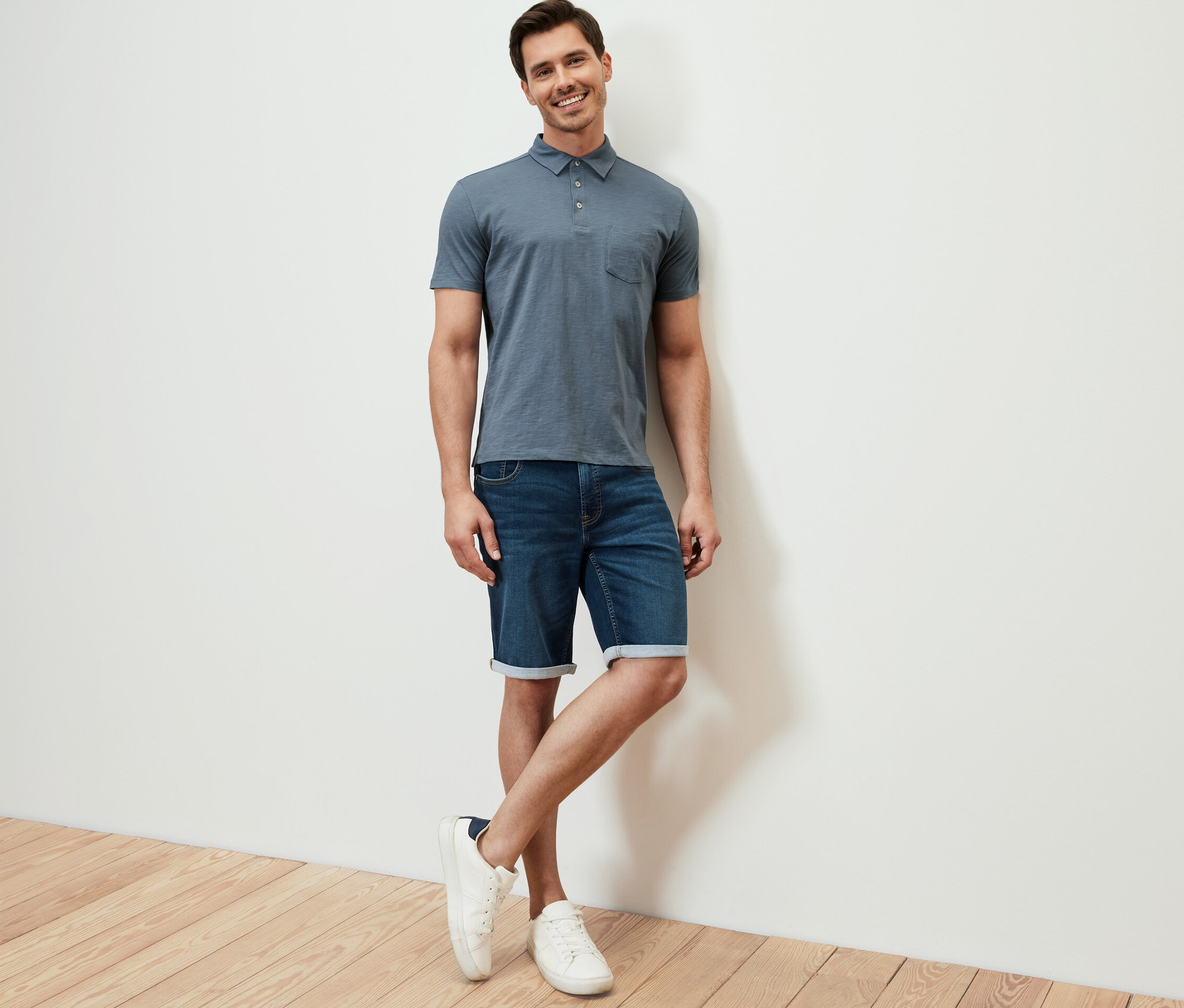 Férfi támaszkodik a falnak, világoskék melírozott jersey pólóinget, farmer rövidnadrágot és fehér tornacipőt visel.