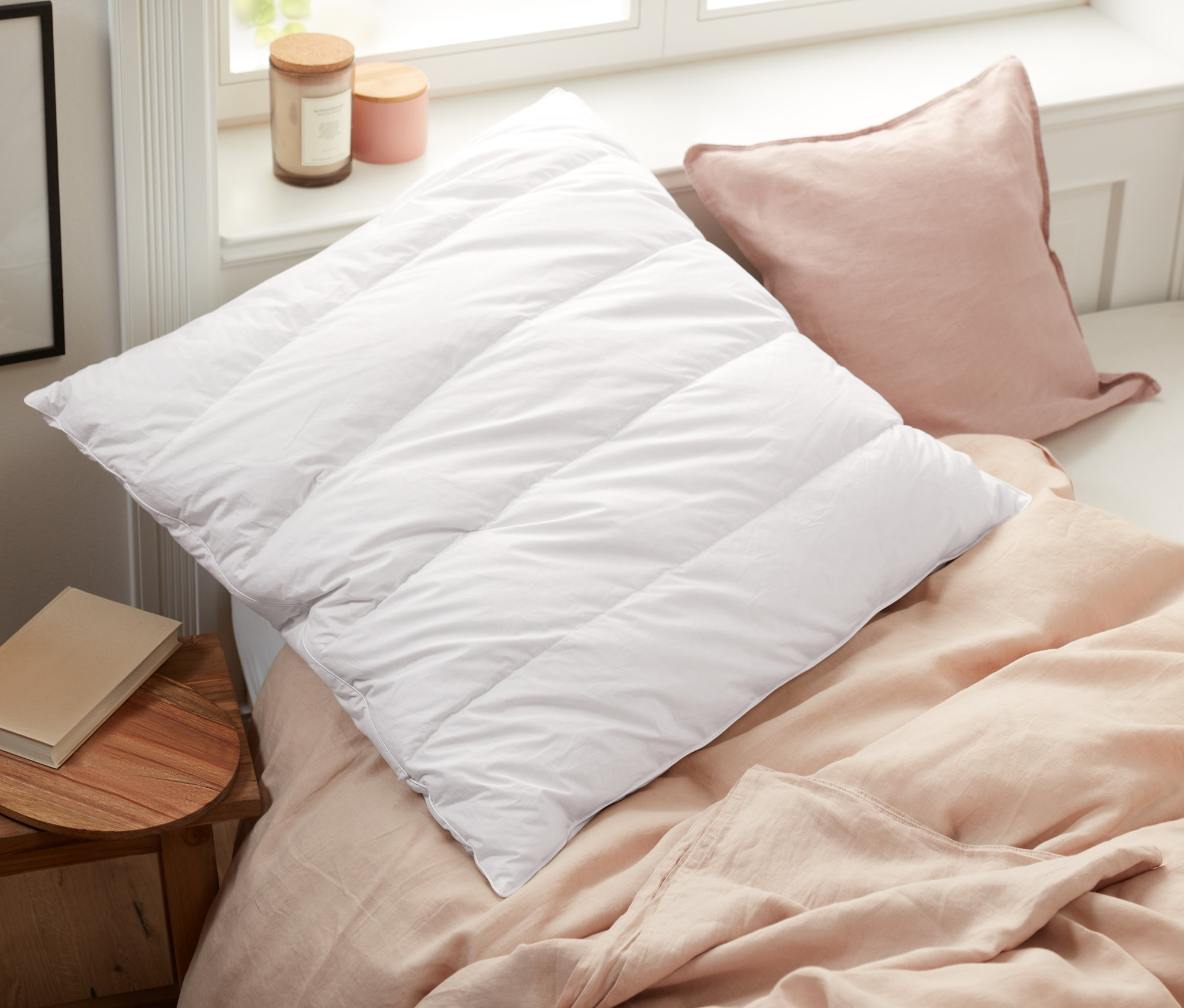 Egy ágyon egy fehér, steppelt párna és egy rózsaszín párna fekszik egy cirbolyafenyő párna mellett.