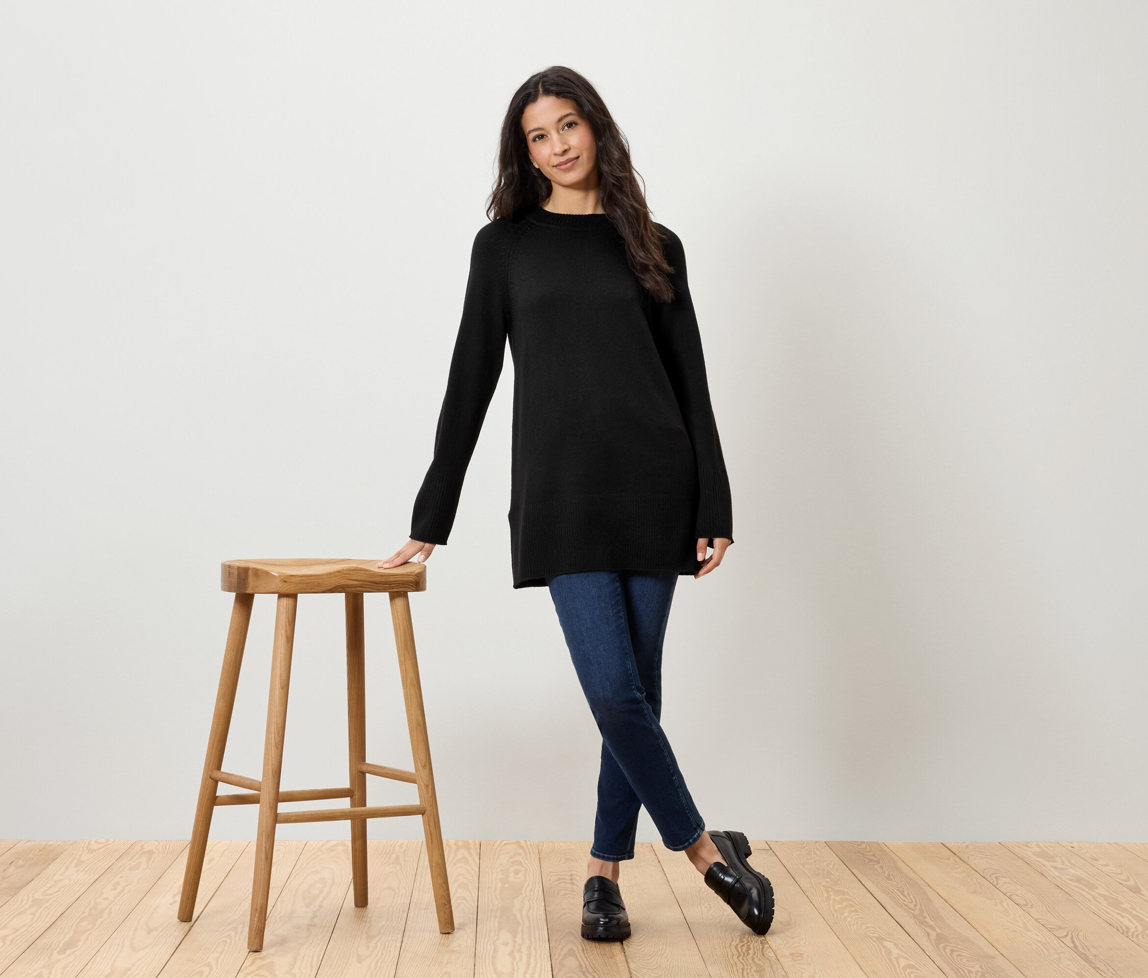Fekete pulóvert és farmert viselő nő áll egy fa szék mellett.