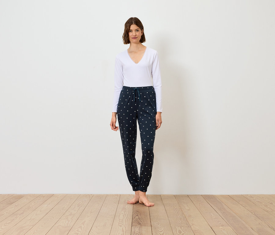 Egy nő áll, fehér hosszú ujjú inget és sötétkék, mintás nadrágot visel.