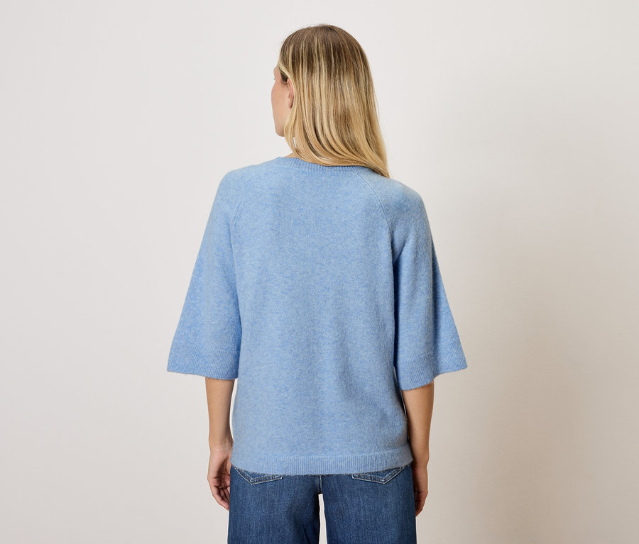 Egy szőke hajú nő áll háttal a kamerának, kék felsőt és farmert visel.