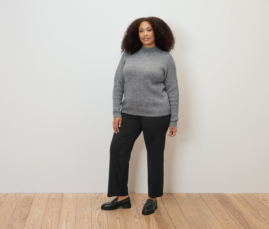 Egy nő áll, szürke, laza kötésű pulóvert és kényelmes jersey nadrágot visel.