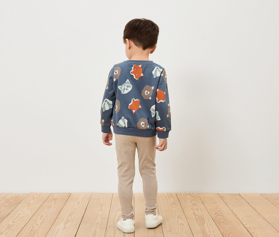 Egy kisfiú háttal áll, kék, állatmintás pulóvert, bézs színű leggingset és fehér cipőt visel.