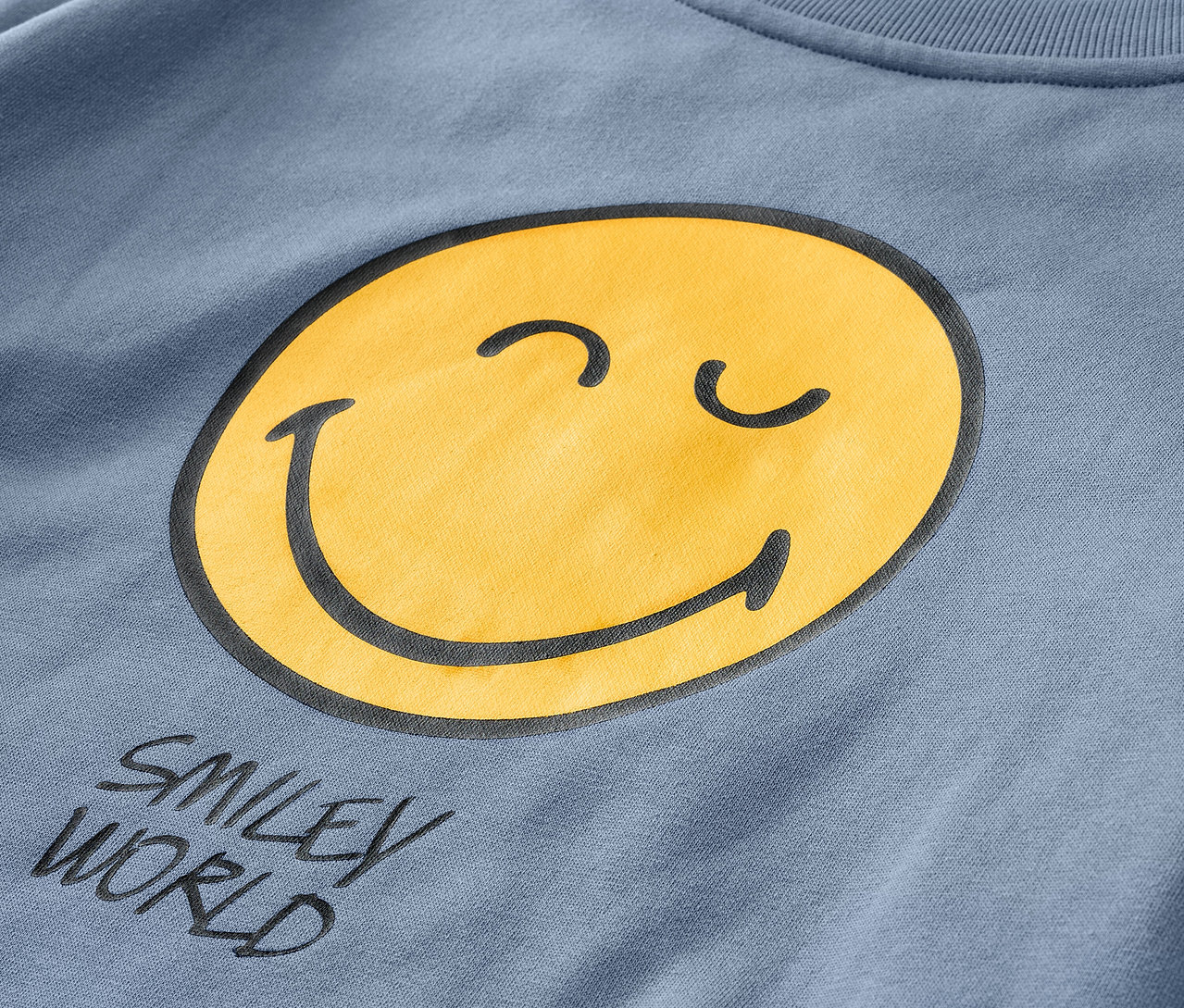 Egy kék SMILEYWORLD® gyerek szabadidőfelső részletfotója, sárga mosolygós fejjel.