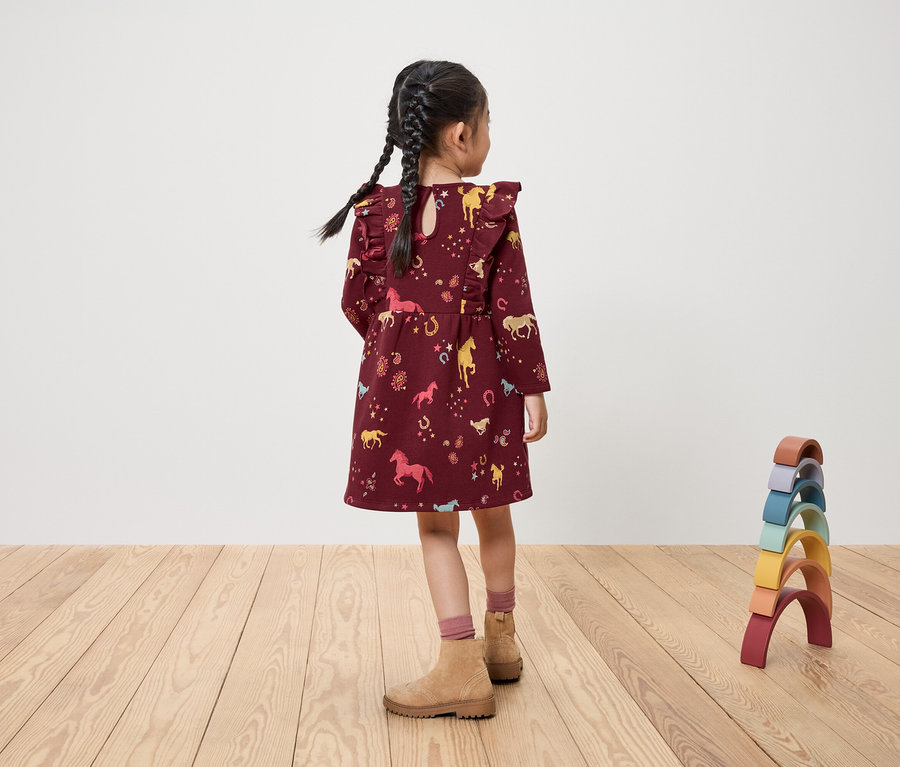 Egy lány háttal áll, bordó, lovas mintás ruhát és játékokat visel.