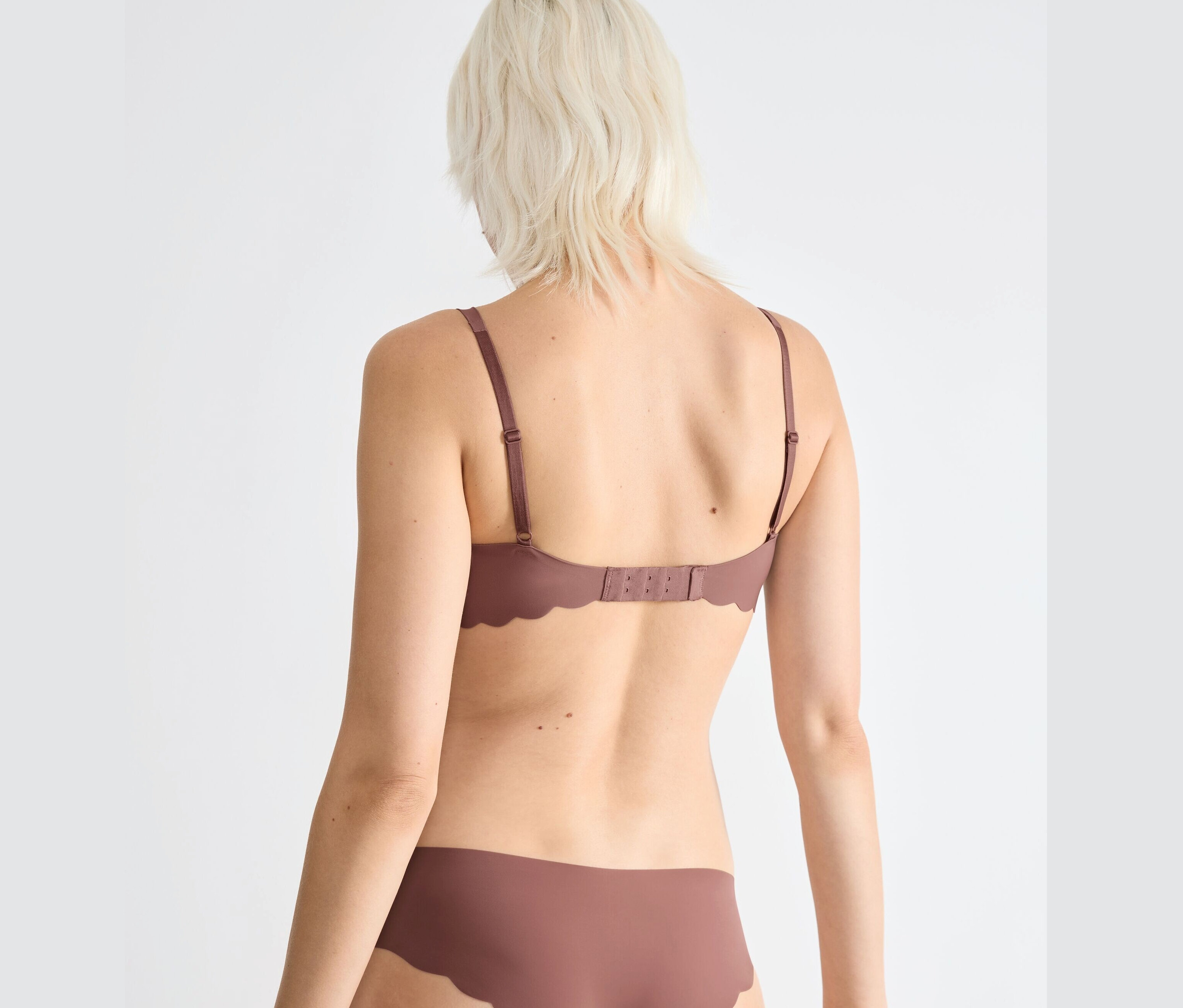 Hátulnézet egy szőke hajú nőről, aki barna sloggi ZERO Microfibre 2.0 puha melltartót és alsót visel.