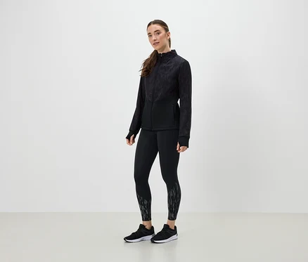 Egy nő fekete sportruházatban pózol világos háttér előtt.